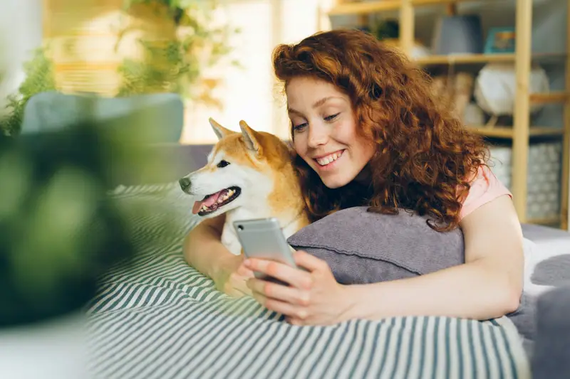 A smiling woman on a sofa looking at her phone, cuddling her dog